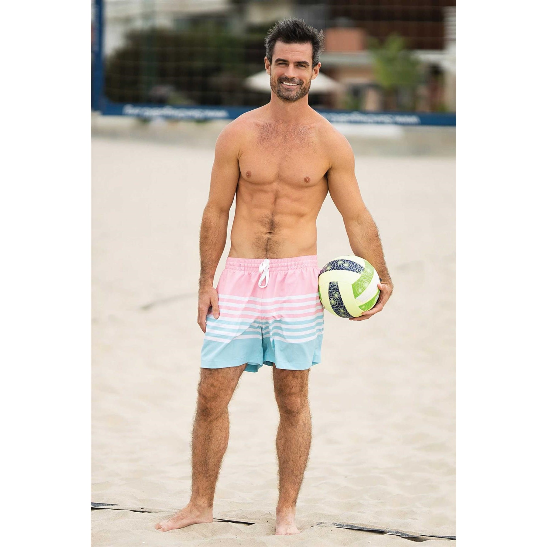 Topless male posed wearing swim trunks on beach holding volleyball