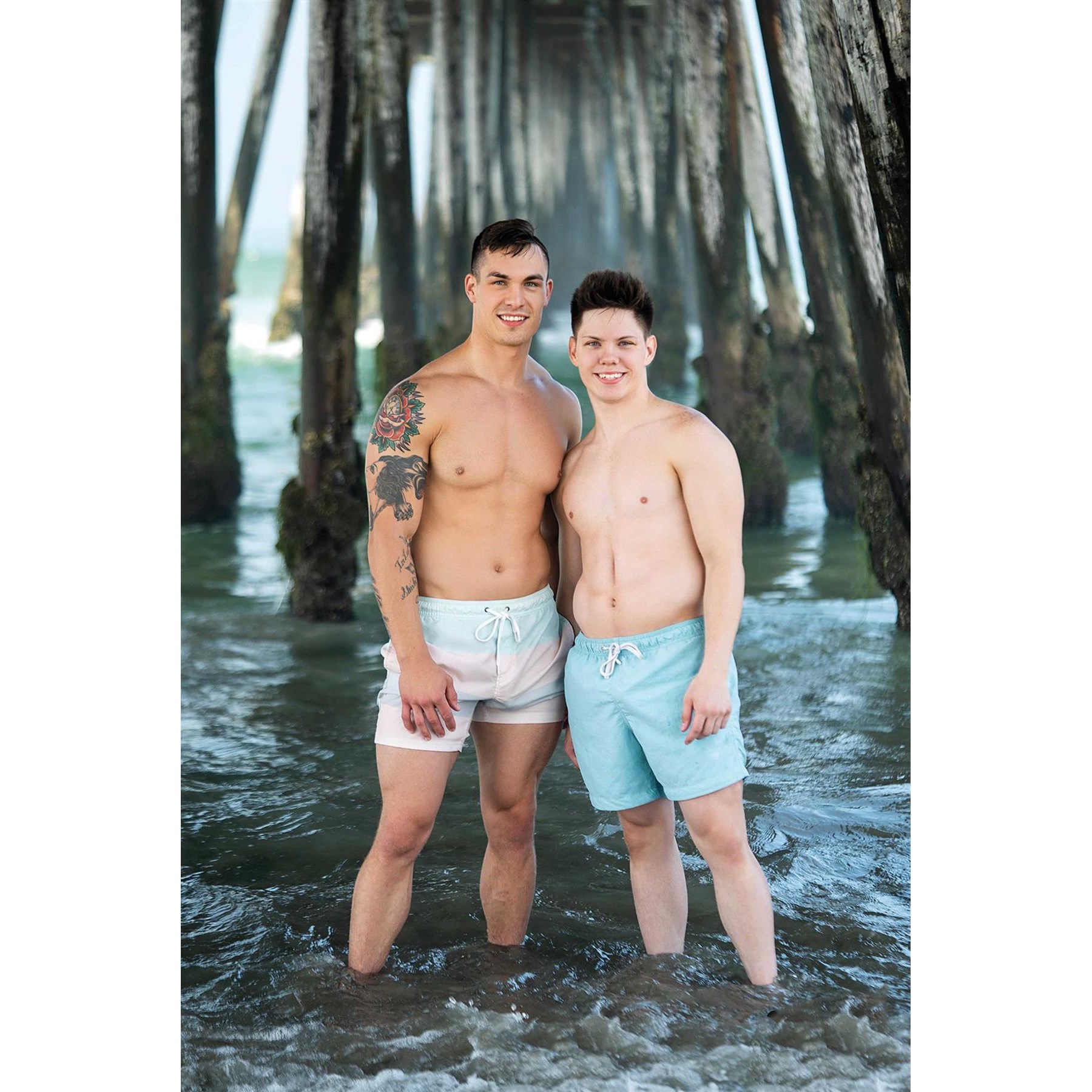 Two topless males posed on beach wearing swim trunks
